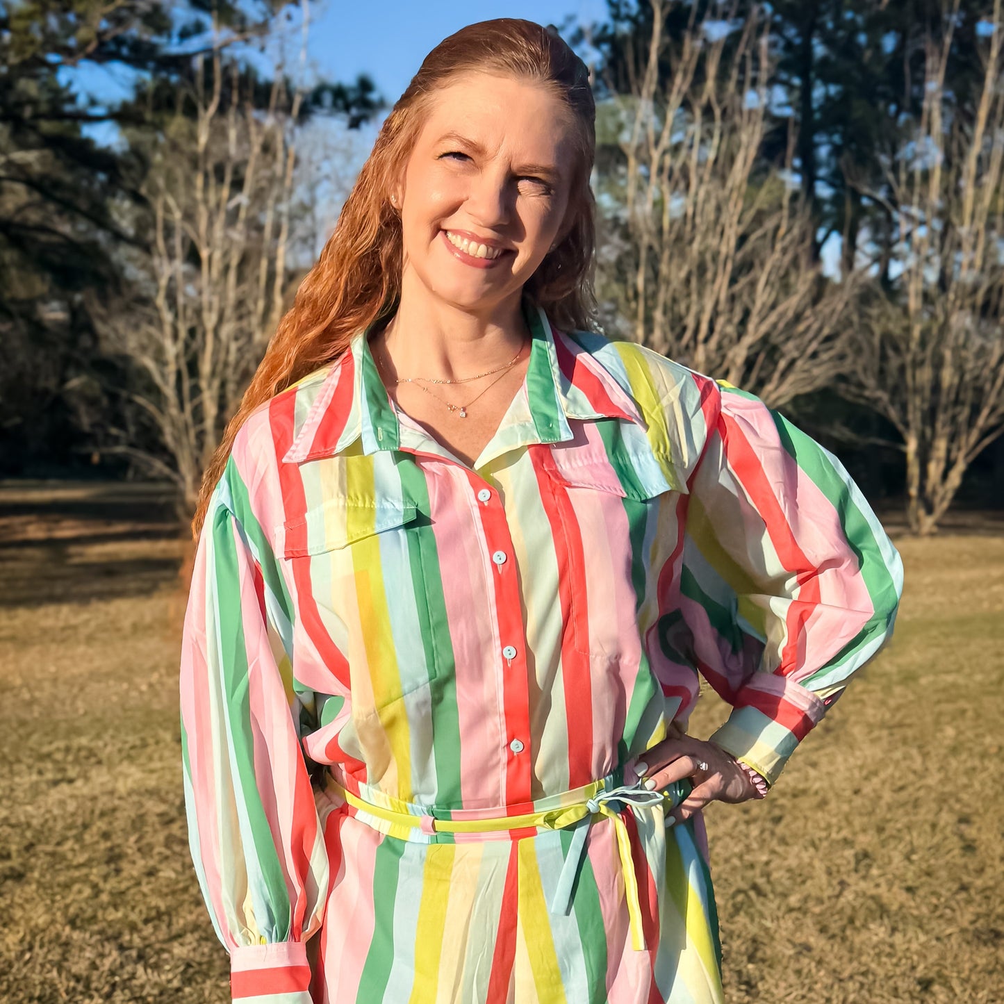 Colorful striped long-sleeve romper with a button-up front, chest pockets, and a matching waist tie, featuring bold vertical stripes in pink, green, yellow, and white.