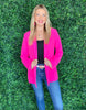 A Woman wearing a Bright Pink Blazer with Bluish Fade Jeans and posing in front of a Green Artificial Grass like Wall.