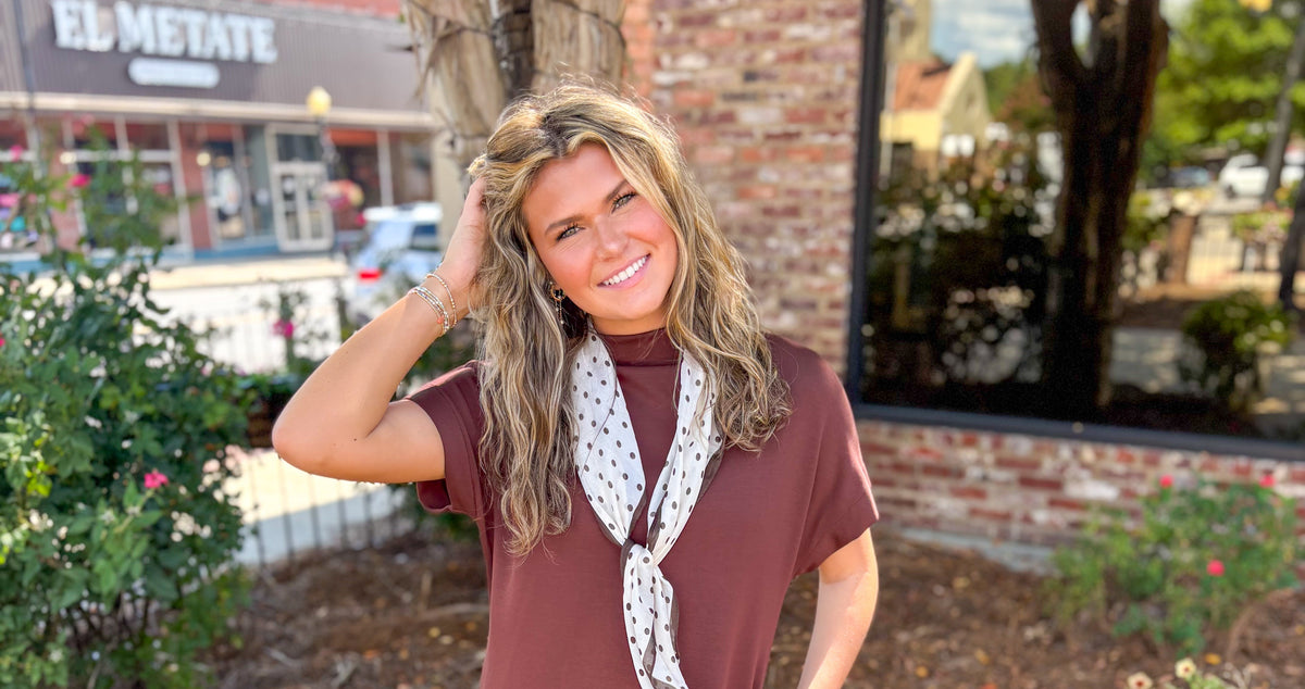 A woman stands outdoors on a sidewalk in front of a brick building, wearing a loose short-sleeve brownish-rose mini dress with a white neck scarf featuring a black polka-dot pattern, and white ankle boots.